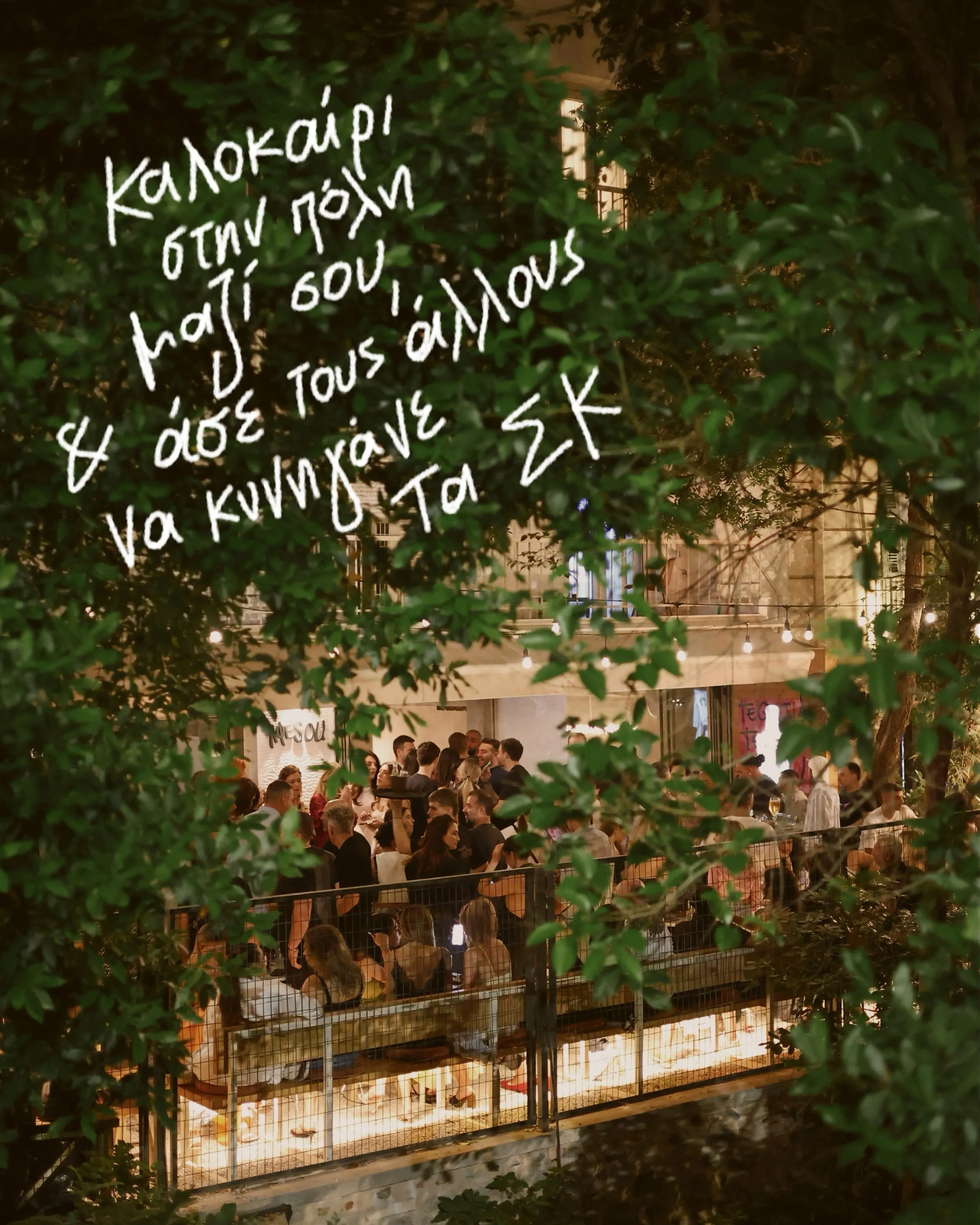 Η φωτισμένη βεράντα της Αφέσου το βράδυ, γεμάτη παρέες, μέσα από τα δέντρα